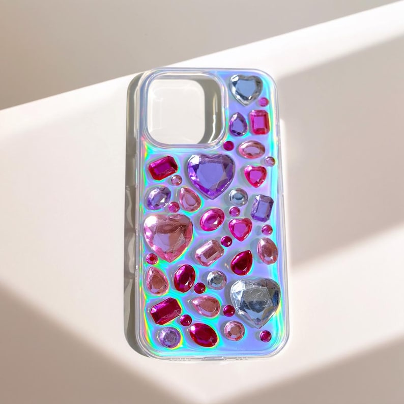 May include: Clear phone case adorned with an array of colorful, iridescent rhinestones. The design features heart, oval, and rectangular shapes in shades of pink, purple, and blue. The case has a clear border and a cutout for the camera.