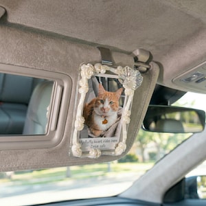 Può includere: Una cornice rettangolare per aletta parasole con un bordo bianco simile a conchiglie contenente la fotografia di un gatto soriano arancione. La parte inferiore della foto presenta il testo stampato: My fluffy heart rides here. Drive safe.