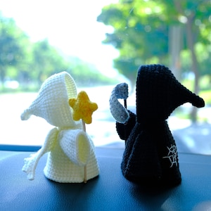May include: Two small crochet figures, one white and one black, both wearing hooded cloaks. The white figure holds a yellow star on a stick, while the black figure holds a light blue scythe. The black figure features a white spiderweb embroidery.
