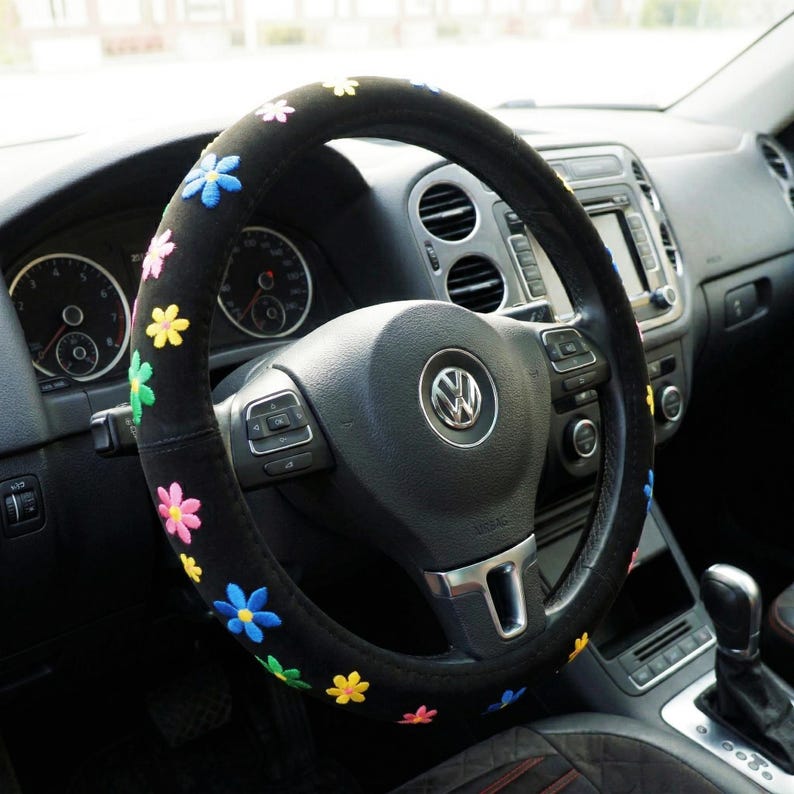 May include: A black steering wheel cover featuring embroidered flowers in various colours. The cover is designed to fit a standard car steering wheel, enhancing the vehicle's interior. The flowers are in shades of pink, yellow, blue, and green.