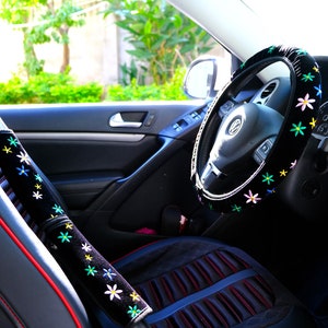 May include: Car interior featuring black accessories with floral embroidery. The steering wheel cover and seatbelt cushions are decorated with colourful flower patterns. The car seat has red stitching.