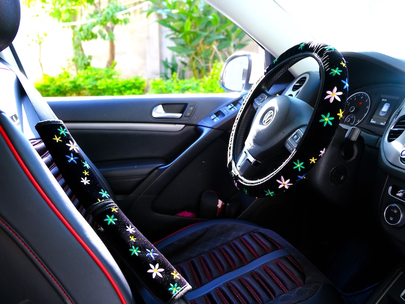 May include: Car interior featuring black accessories with floral embroidery. The steering wheel cover and seatbelt cushions are decorated with colourful flower patterns. The car seat has red stitching.