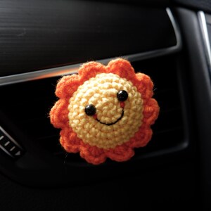 Puede incluir: Un clip de ventilaci&oacute;n de coche con forma de sol tejido a ganchillo. El sol tiene un centro amarillo con un borde naranja con volantes. Cuenta con ojos de bot&oacute;n negros y una sonrisa cosida. El clip est&aacute; dise&ntilde;ado para sujetarse a una rejilla de ventilaci&oacute;n del coche.