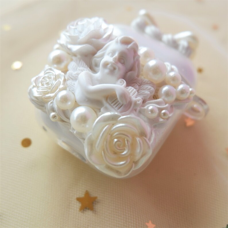 May include: A white, pearlescent trinket box with a sculpted cherub, roses, and faux pearls. The box has a square shape with rounded edges and a bow detail. The cherub is playing a harp.