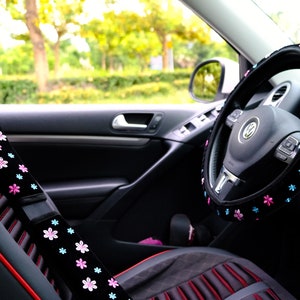 May include: Black car interior with a black steering wheel cover with pink and blue flowers. The steering wheel cover is made of soft, plush fabric. The car seat is black with red stitching.