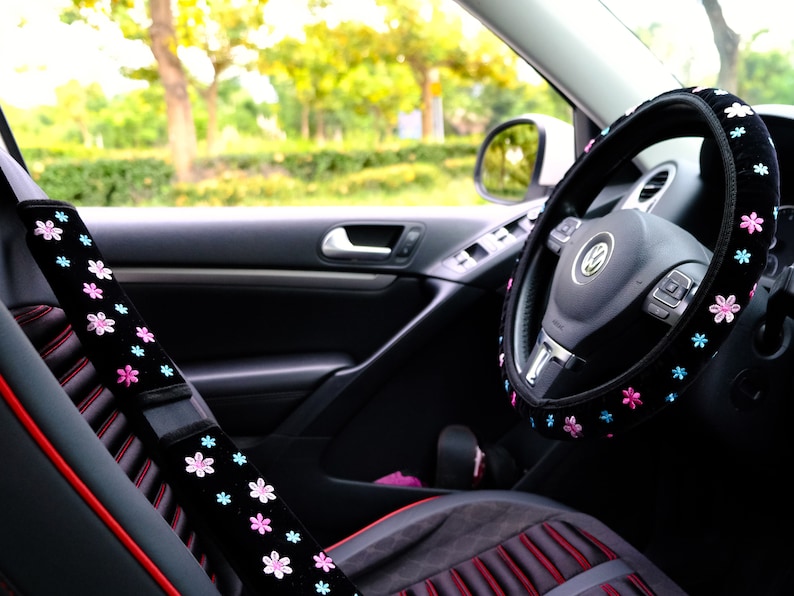 May include: Black car interior with a black steering wheel cover with pink and blue flowers. The steering wheel cover is made of soft, plush fabric. The car seat is black with red stitching.