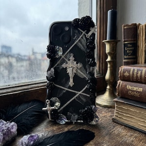 May include: Black phone case with a gothic aesthetic. The case features a silver cross pendant, black roses, and decorative chains. The case is embellished with clear crystals and a marble-like pattern. The phone case is a dark and elegant accessory.
