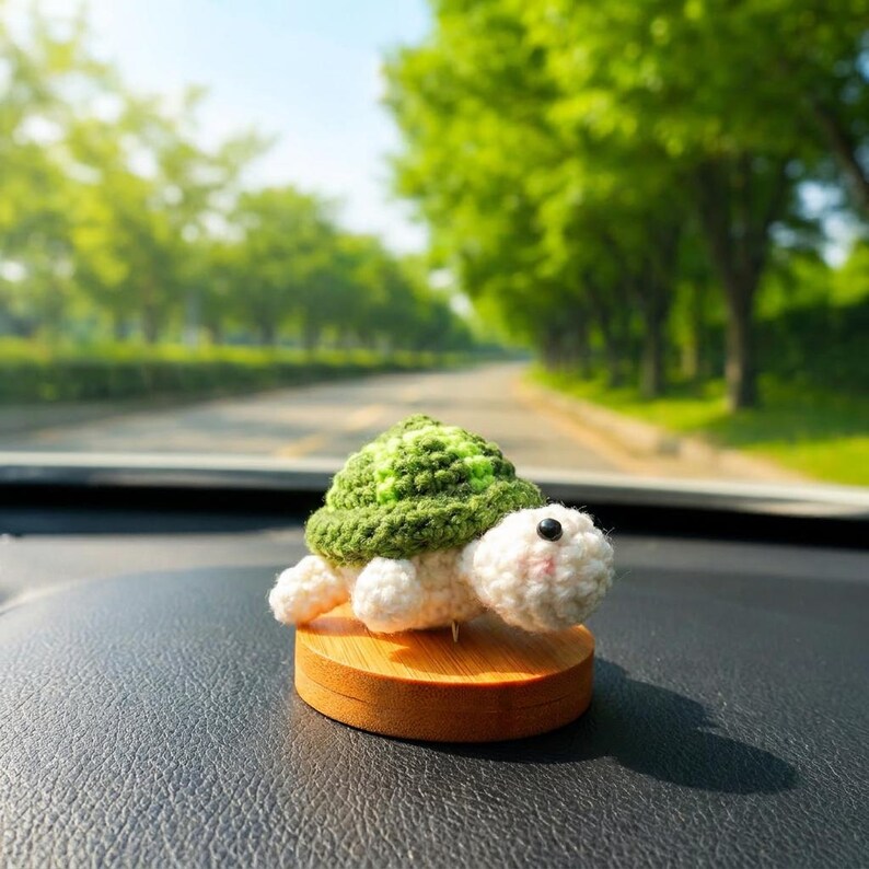 May include: A crocheted turtle figurine with a green shell and white body sits on a wooden base. The turtle has a smiling face and black eyes. The background shows a road and trees.