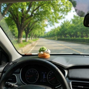 May include: A crocheted turtle figurine with a green shell and white body sits on a wooden base on a car dashboard. The car's steering wheel and a road lined with trees are visible through the windshield.