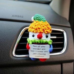 May include: A handmade crochet clown car air vent decoration. The clown has a white face, a red nose, and a yellow and green hat. It holds a sign that reads, "Not all who wander are lost—some are just chasing confetti!"