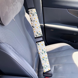 May include: Two rectangular seat belt covers featuring a white background with an embroidered pattern of small white daisy flowers with yellow centers and green stems. The covers are secured around a gray seat belt in a vehicle interior.
