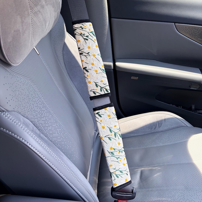 May include: Two rectangular seat belt covers featuring a white background with an embroidered pattern of small white daisy flowers with yellow centers and green stems. The covers are secured around a gray seat belt in a vehicle interior.
