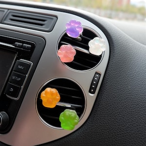 Könnte beinhalten: Eine Auswahl an pfotenförmigen Auto-Lufterfrischern in den Farben Lila, Rosa, Weiß, Orange und Grün. Diese dekorativen Lufterfrischer werden in die Lüftungsschlitze eines Autos gesteckt und werten den Innenraum des Fahrzeugs optisch und geruchlich auf.