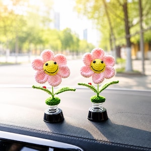 May include: Two crochet flower car decorations. Each flower has pink petals, a yellow smiling face, and green leaves. The flowers are attached to a black base with a spring. A red ladybird sits on one of the leaves.