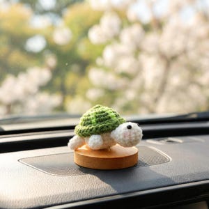 May include: A crocheted turtle ornament with a green shell and white body, set on a small, round wooden base. The turtle has black eyes and a pink blush. The ornament is displayed on a car dashboard.