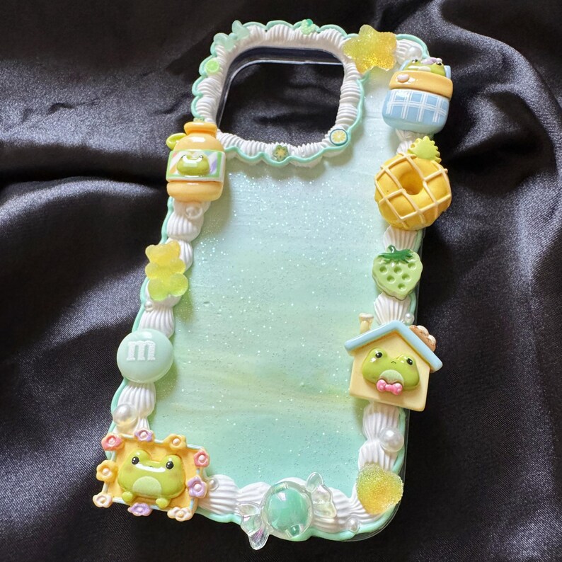 May include: A decorative phone case with a light green and white background, adorned with various 3D embellishments. These include a frog in a house, a yellow donut, a strawberry, and a jar. The case is edged with a white, textured border.