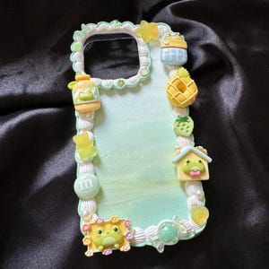 May include: A decorative phone case with a light green and white background, adorned with various miniature food and frog-themed embellishments. Includes a yellow pineapple, strawberry, and a frog house. The case is edged with a white, textured border.