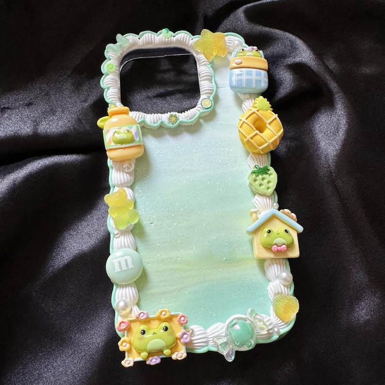 May include: A decorative phone case with a light green and white background, adorned with various miniature food and frog-themed embellishments. Includes a yellow pineapple, strawberry, and a frog house. The case is edged with a white, textured border.