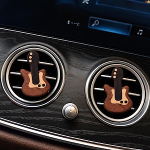 May include: Two wooden guitar-shaped car air freshener clips attached to the circular silver vents of a vehicle dashboard. The guitar ornaments feature a dark brown neck, a lighter brown body, and small circular accents on the headstock and body.