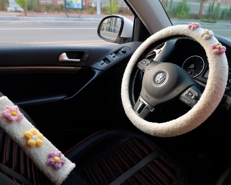 May include: White faux fur steering wheel cover with pink flower embellishments. The cover is installed on a black leather steering wheel with a silver Volkswagen logo.