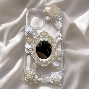 May include: A clear phone case decorated with white and translucent cream-coloured floral embellishments, small pearls, and clear butterfly accents. Centered on the back is an oval-shaped, ornate cream-coloured frame surrounding a small mirror.