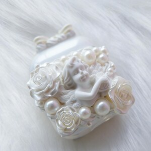May include: A white, heart-shaped decorative object with a cherub, roses, and pearls. The cherub is playing a harp. The roses and pearls are arranged around the cherub. The object has a pearlescent finish.