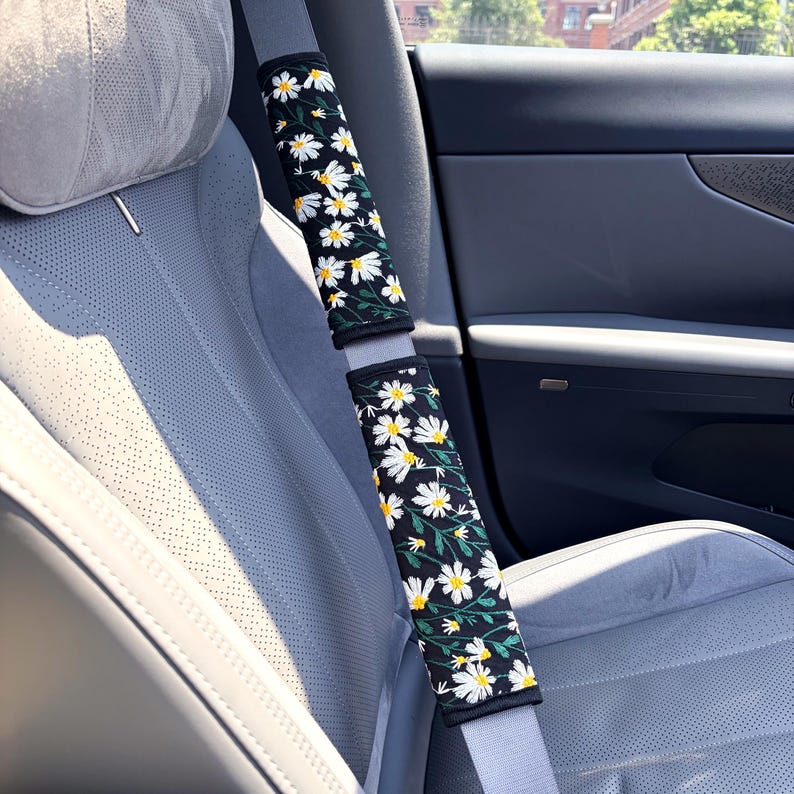 May include: Two rectangular seat belt covers featuring a black fabric base with a repeating pattern of white daisy flowers, yellow centers, and green stems. The covers are wrapped around a light gray seat belt inside a vehicle interior.