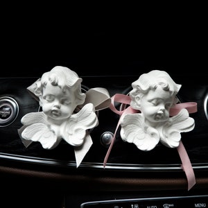 May include: Two white porcelain angel figurines with wings, placed on a black dashboard of a car. The angels have pink and white ribbons attached to them.