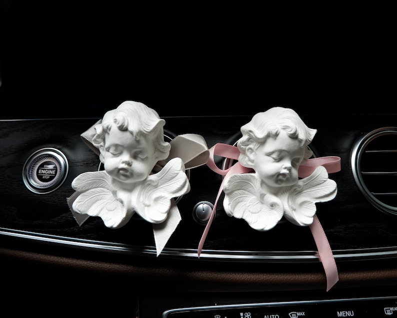 May include: Two white porcelain angel figurines with wings, placed on a black dashboard of a car. The angels have pink and white ribbons attached to them.