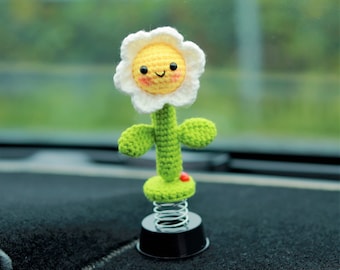 Könnte beinhalten: Eine gehäkelte Blume mit einem lächelnden Gesicht, befestigt an einer Feder und einem schwarzen Sockel. Die Blume ist weiß mit einer gelben Mitte und grünen Blättern.