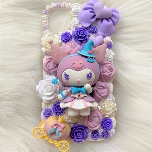 Puede incluir: Una funda de tel&eacute;fono blanca decorada con flores 3D de color morado, blanco y rosa. Un personaje de dibujos animados morado y blanco con un sombrero de bruja est&aacute; en el centro de la funda. El personaje sostiene una varita con una estrella en el extremo. Un carruaje dorado con un lazo rosa y blanco est&aacute; en la parte inferior de la funda.