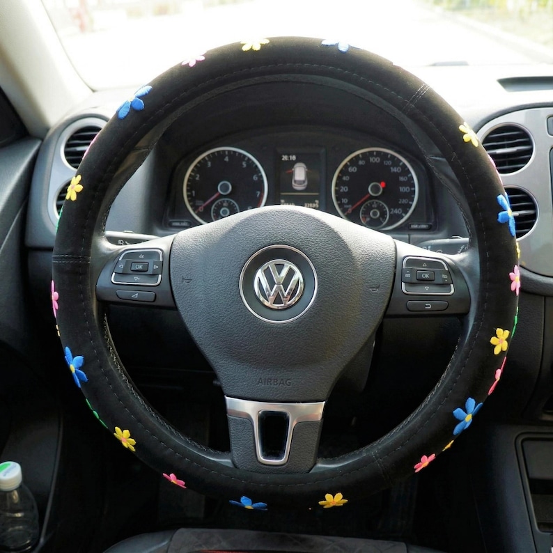 May include: Black steering wheel cover with embroidered colourful flowers. The cover is on a Volkswagen steering wheel. The flowers are yellow, pink, and blue. The car's dashboard and gauges are visible in the background.