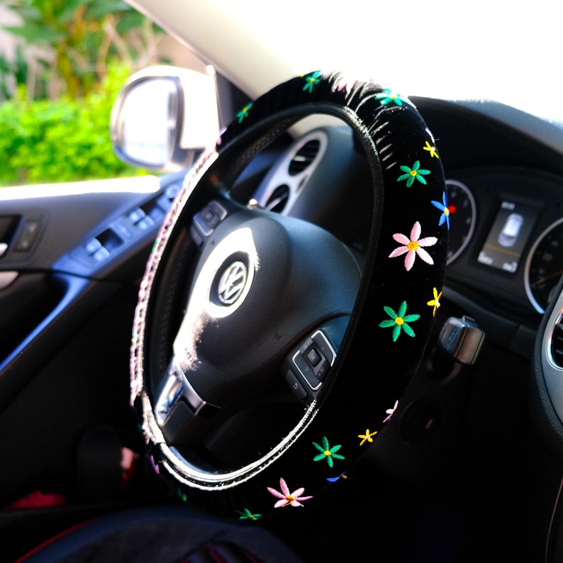 May include: A black steering wheel cover adorned with embroidered flowers in various colours. The cover is fitted on a black steering wheel within a car's interior. The flowers are pink, yellow, blue, and green. The car's dashboard is also visible.