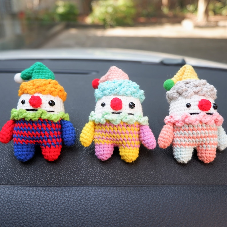 May include: Three crocheted clown dolls with different coloured outfits. The first doll is wearing a red and blue checked outfit with a green hat. The second doll is wearing a pink and yellow striped outfit with a blue hat. The third doll is wearing a peach and white striped outfit with a grey hat.