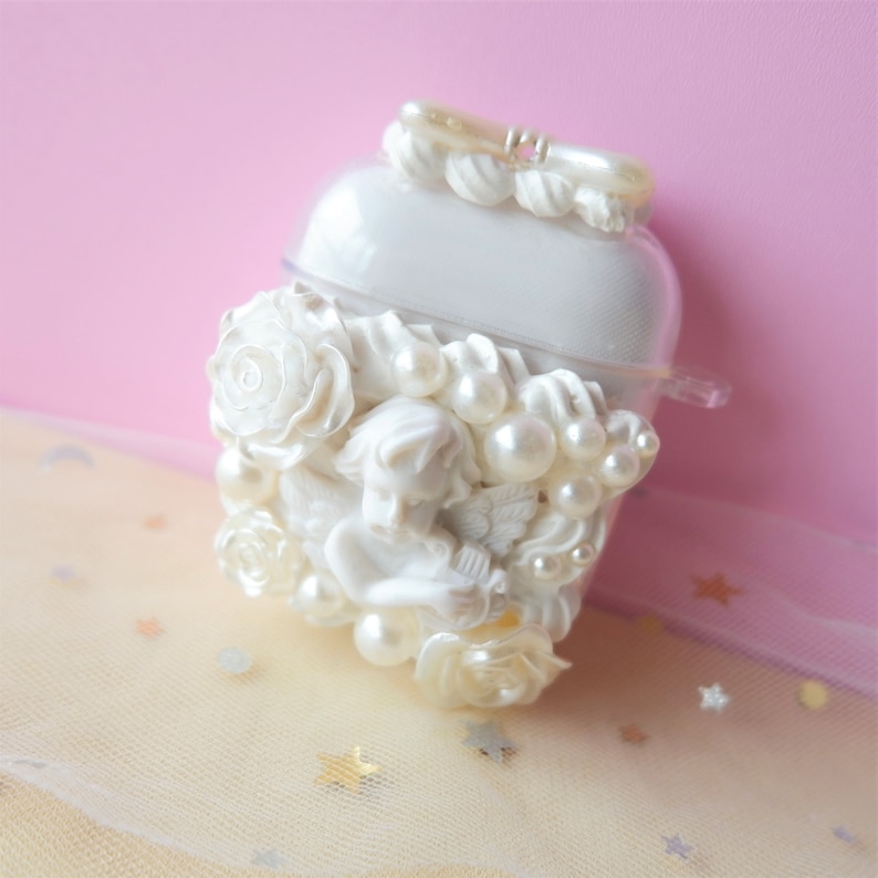 May include: A white, decorative case with a sculpted cherub, roses, and faux pearls. The case has a clear plastic top and a small handle. The design is ornate and detailed, with a romantic, vintage aesthetic.