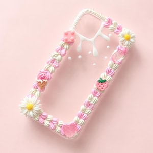 May include: A clear phone case decorated with white and pink whipped cream-style piping along the edges. The design features small charms including a pink building block, a daisy, a strawberry, an ice cream cone, and a pink heart.