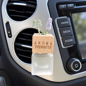 Könnte beinhalten: Ein Auto-Lufterfrischer mit einem quadratischen Flakon aus Milchglas, der mit einem hellen Holzblock verschlossen ist, in den der Text AROMA THERAPIE eingraviert ist. Zwei kleine gehäkelte Blumenstiele, einer hellgrün und einer helllila, ragen aus dem Holz.