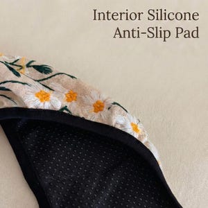 May include: A beige fabric headband featuring embroidered white daisy flowers with yellow centers and green stems. The underside is a black interior silicone anti-slip pad with a textured dotted surface to provide grip during wear.