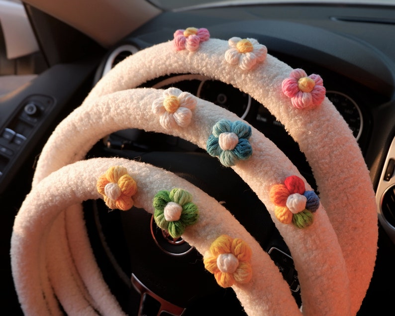May include: A white fuzzy steering wheel cover with colorful crocheted flowers attached. The flowers are in shades of pink, blue, green, yellow, and red.