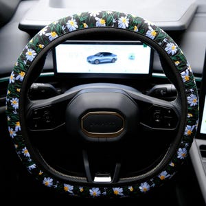 May include: A black steering wheel cover featuring an embroidered pattern of white daisy flowers with yellow centers and green stems. The cover is fitted over a black steering wheel with a central horn button and integrated control buttons.