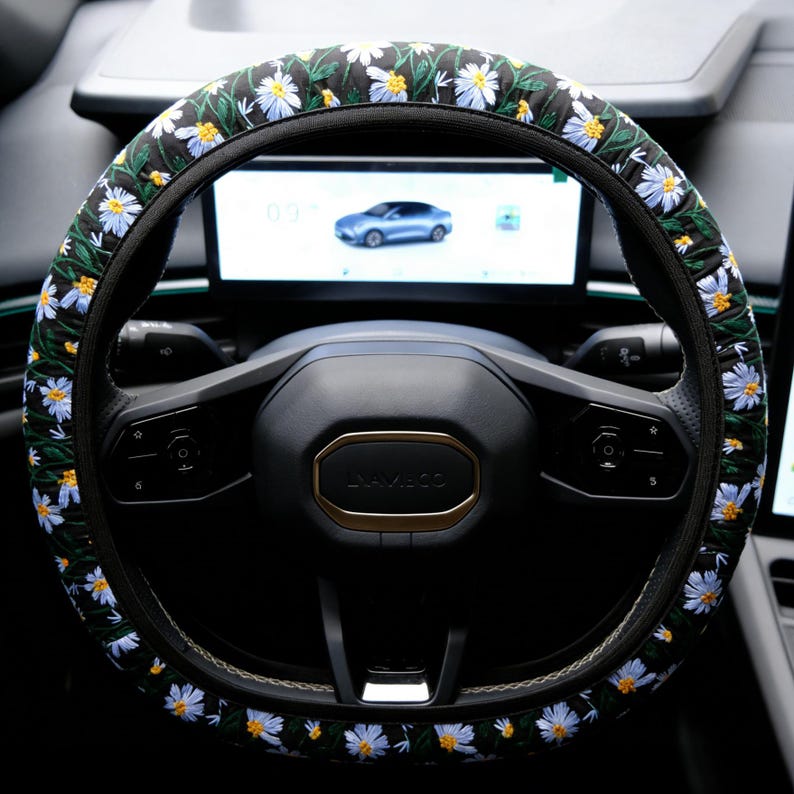 May include: A black steering wheel cover featuring an embroidered pattern of white daisy flowers with yellow centers and green stems. The cover is fitted over a black steering wheel with a central horn button and integrated control buttons.