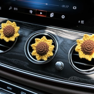May include: Three crocheted sunflower car air fresheners in yellow and brown. The sunflowers are placed in the air vents of a car dashboard.