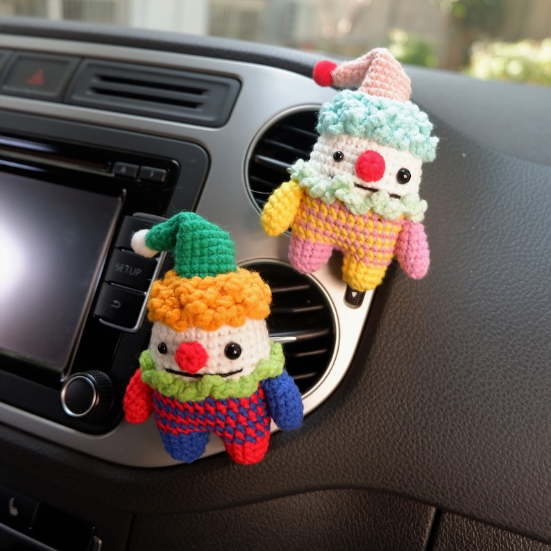 May include: Two crocheted clown dolls with colourful hats and outfits hanging from the air vent of a car. The clown on the left has a green hat and a red, blue and yellow outfit. The clown on the right has a pink hat and a pink, yellow and blue outfit.