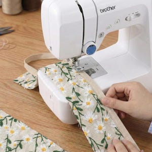 May include: A white Brother sewing machine stitching a beige fabric strip featuring an embroidered pattern of white daisy flowers with yellow centers and green stems. The machine has a blue dial and a metal needle plate.