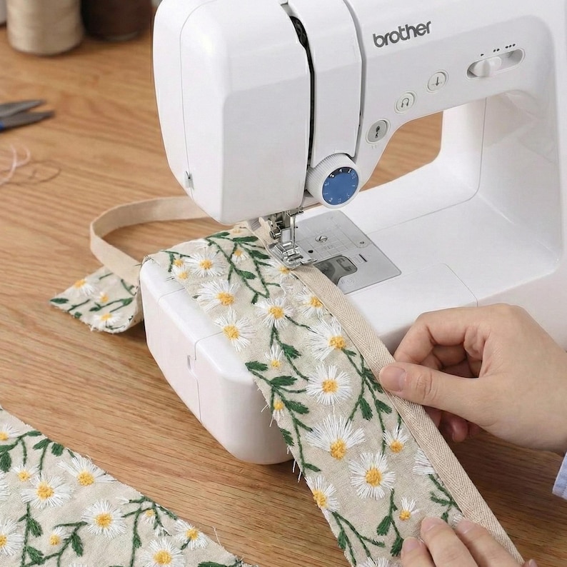 May include: A white Brother sewing machine stitching a beige fabric strip featuring an embroidered pattern of white daisy flowers with yellow centers and green stems. The machine has a blue dial and a metal needle plate.