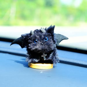 May include: A plush black creature with blue eyes, small horns, and bat-like wings. It wears a silver cross pendant and sits on a small wooden base. The toy is designed to resemble a devilish animal.