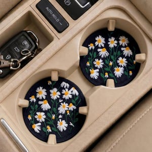 May include: Two navy blue car coasters with embroidered white daisies and yellow centers. The coasters are placed in a car's cup holders, next to a car key and the "AUTO HOLD" button.