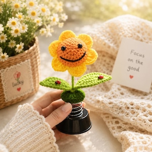 May include: A crocheted sunflower with a smiling face, yellow petals, and an orange centre. The green leaves have red ladybug accents. The base is black with a silver spring. A card reads "Focus on the good".