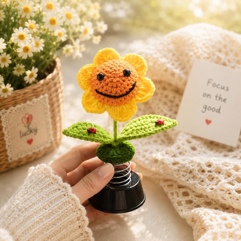 May include: A crocheted sunflower with a smiling face, yellow petals, and an orange centre. The green leaves have red ladybug accents. The base is black with a silver spring. A card reads "Focus on the good".