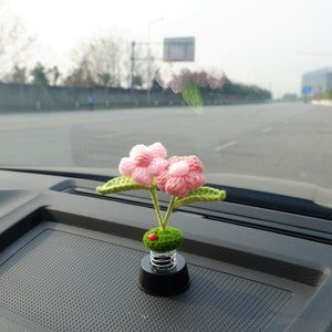 May include: A pink and green crocheted flower car air freshener with a spring base. The air freshener is sitting on the dashboard of a car.